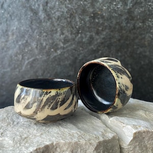 Può includere: Due piccole ciotole in ceramica con un interno nero profondo e un esterno a contrasto caratterizzato da un motivo marmorizzato con pennellate color crema e marrone scuro. Le ciotole hanno una forma arrotondata e un bordo spesso.