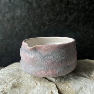 Może przedstawiać: Mała ceramiczna miseczka o teksturowanej, matowej powierzchni zewnętrznej w nakrapianym odcieniu delikatnego różu i łupkowego szarości. Wnętrze jest gładkie, wykończone błyszczącą białą glazurą, z subtelnym dzióbkiem na górnej krawędzi.