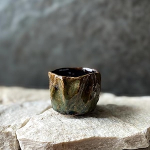 Może przedstawiać: Mały, ręcznie formowany kubek ceramiczny z głęboką, błyszczącą glazurą w odcieniach mchu, brązu i ciemnego grafitu. Zewnętrzna powierzchnia posiada nieregularne, pofalowane wgłębienia oraz gruby, nierówny brzeg, co nadaje jej teksturalny wygląd.