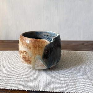 Może przedstawiać: Mała, okrągła ceramiczna miseczka z szerokim otworem i subtelnym dzióbkiem. Zewnętrzna strona pokryta jest reaktywnym szkliwem w odcieniach ciepłego bursztynu, głębokiego brązu i przygaszonego błękitu, tworząc płynny, marmurkowy wzór na powierzchni.