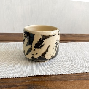 Może przedstawiać: Ceramiczny kubek o kremowej podstawie z wyrazistymi, zamaszystymi czarnymi pociągnięciami pędzla owijającymi się wokół zewnętrznej strony. Naczynie ma zaokrągloną sylwetkę bez ucha, z gładką, otwartą krawędzią i solidną konstrukcją.