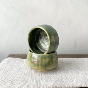 Puede incluir: Dos cuencos de cerámica apilados con un esmalte verde oliva brillante y moteado con texturas acanaladas verticales. El interior del cuenco superior es oscuro y reflectante, mientras que el exterior muestra variaciones en tonos verdes.