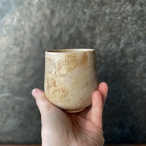 Może przedstawiać: Mały, bezuchy kubek ceramiczny o zaokrąglonym, pękatym kształcie. Zewnętrzna powierzchnia ma teksturowane, nakrapiane wykończenie w odcieniach kremu, beżu i jasnego brązu, z widocznymi różnicami w szkliwie, które nadają mu rustykalny wygląd.