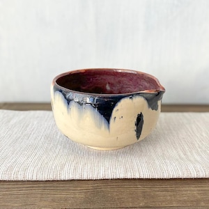 Peut inclure: Un bol en céramique de forme arrondie avec un petit bec verseur. L'intérieur présente une glaçure violet prune profond, tandis que l'extérieur affiche une base couleur crème avec des coulures bleu marine foncé descendant du bord.