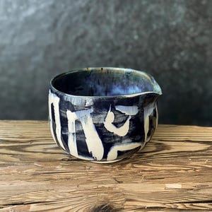 Op de afbeelding: Een keramische kom met een schenktuit, voorzien van een donker marineblauw glazuur en gedurfde, met de hand geschilderde witte penseelstreekpatronen. De binnenkant van de kom heeft een diepblauwe en zwarte afwerking met een gladde textuur.