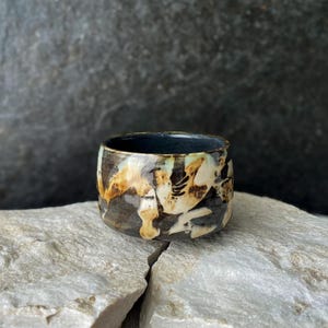 Może przedstawiać: Mały, okrągły kubek ceramiczny z ciemnym, błyszczącym wnętrzem oraz powierzchnią zewnętrzną ozdobioną abstrakcyjnym, cętkowanym wzorem kremowych, beżowych i ciemnobrązowych szkliw, które tworzą warstwowy, organiczny wygląd na korpusie.