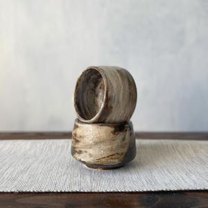 Op de afbeelding: Twee handgemaakte keramische bekers, de ene op de andere gestapeld. De bekers vertonen een rustieke, aardse esthetiek met beige, bruine en grijze tinten. Het getextureerde oppervlak en de unieke vorm suggereren een ambachtelijke oorsprong.