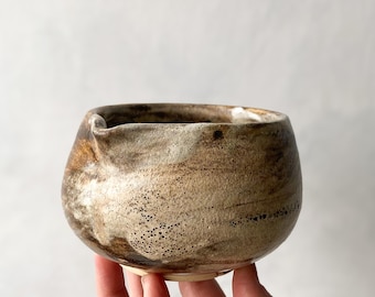 May include: A small, round ceramic bowl with a textured, mottled surface in shades of beige, brown, and dark gray. The vessel features a distinct, slightly protruding pouring spout on one side and a wide, open rim with a thick, uneven edge.