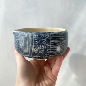 Peut inclure: Un bol en céramique avec un bec verseur, présentant un extérieur bleu profond orné de motifs blancs gravés, notamment des lignes verticales, de petites fleurs et des hachures. L'intérieur du bol a une finition émaillée blanc cassé lisse.