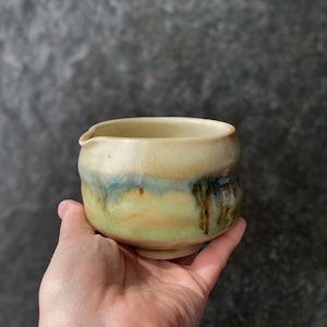 Pode incluir: Uma pequena tigela de cerâmica para servir com formato arredondado e esmalte base bege claro. O exterior apresenta uma faixa horizontal de tons azuis e verdes misturados, com detalhes de esmalte castanho escuro em forma de gotas perto do bico.