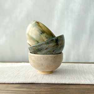 May include: A stack of three ceramic bowls. The top bowl is cream with green and brown streaks. The middle bowl is blue-grey with dark speckles. The bottom bowl is a solid, textured light beige with a matte finish and a small circular base.