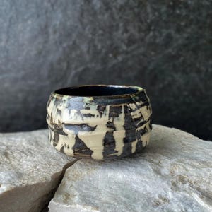 Könnte beinhalten: Eine kleine, runde Keramikschale mit einem dunkel glänzenden schwarzen Inneren und einer texturierten Außenseite mit einem kontrastierenden Tropfmuster aus cremefarbener Glasur auf dunkelbraunem Grund und einer horizontalen Kante.