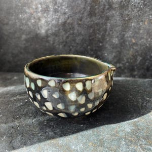 Peut inclure: Un petit bol en céramique rond avec un vernis sombre et brillant présentant un motif de taches irrégulières de couleur blanc cassé. Le bol possède un bord lisse et incurvé avec un bec verseur intégré discret sur un côté et un intérieur profond.