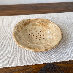 Può includere: Un portasapone rotondo in ceramica beige con una superficie irregolare e testurizzata e una serie di piccoli fori di drenaggio circolari raggruppati al centro. Il piatto presenta tonalità marroni e beige screziate lungo il bordo irregolare.