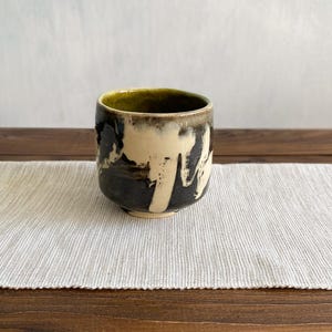 Może przedstawiać: Ceramiczny kubek o zaokrąglonym kształcie z wykończeniem z reaktywnego szkliwa. Zewnętrzna strona posiada kontrastowy wzór z kremowymi zaciekami na ciemnoszarym tle, a wnętrze pokryte jest gładkim, oliwkowozielonym szkliwem.