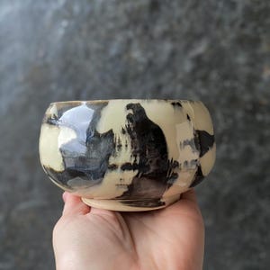 Pode incluir: Uma pequena tigela de cerâmica arredondada com acabamento brilhante e um padrão marmorizado de esmalte base creme com pinceladas pretas ousadas e amplas. O recipiente tem uma silhueta curva suave e uma base circular definida.
