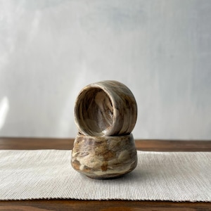 Pode incluir: Duas tigelas de cerâmica feitas à mão empilhadas. As tigelas têm uma aparência marmorizada com tons de castanho, bege e creme. O interior das tigelas é castanho mais escuro. As tigelas estão sobre um caminho de mesa branco. Cada tigela tem cerca de 8 cm de diâmetro.