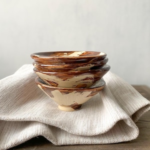 Pode incluir: Uma pilha de três pequenas tigelas de cerâmica com uma base de cor creme e padrões irregulares de pinceladas castanhas pintadas à mão. Cada tigela tem uma borda larga e alargada e uma base circular estreita com um acabamento brilhante.