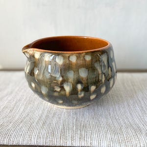Op de afbeelding: Een keramieken melkkannetje met een rond lichaam en een kleine schenktuit. De buitenkant heeft een donkerbruin en grijs glazuur met een patroon van lichte, cirkelvormige witte stippen. De binnenkant van de kan heeft een gladde, oranjebruine afwerking.