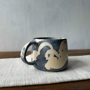 Könnte beinhalten: Ein Keramikbecher mit handbemaltem Design in dunkelanthrazitfarbener, texturierter Ausführung, kontrastiert durch organische, fließende cremefarbene Formen. Der Becher hat einen abgerundeten Körper und einen stabilen, geschwungenen Henkel.