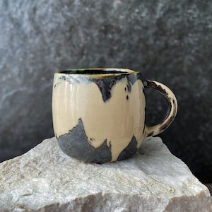 Può includere: Una tazza in ceramica con corpo arrotondato e manico laterale. L'esterno presenta uno smalto bicolore con una sezione centrale liscia color beige chiaro che contrasta con uno smalto grigio antracite scuro e testurizzato che cola dal bordo.