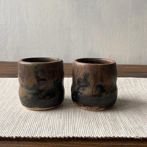Può includere: Due piccole tazze cilindriche in ceramica caratterizzate da una smaltatura scura e maculata con toni misti di marrone profondo, nero e accenni di verde spento. La superficie presenta una finitura liscia e lucida con sottili variazioni di colore.