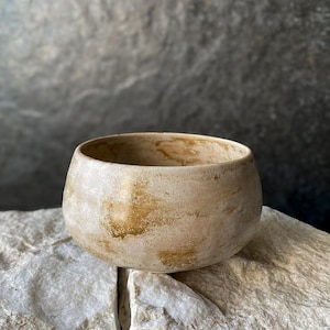 May include: A round ceramic bowl with a textured surface featuring a blend of cream, beige, and golden-brown tones. The exterior has a mottled, weathered appearance with visible brushstrokes and a matte finish, creating a natural, earthy aesthetic.
