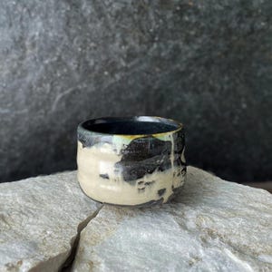 Pode incluir: Uma pequena chávena de cerâmica redonda com um esmalte reativo que apresenta bandas contrastantes em tons de creme e preto carvão escuro. A superfície tem um acabamento liso e brilhante com gotas visíveis e uma mistura orgânica de cores.