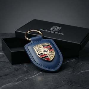 May include: Blue leather Porsche keychain with a gold-colored ring. The keychain features the Porsche emblem in gold, red, black, and white. A black box with the Porsche logo is in the background.