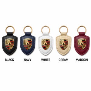 May include: Five shield-shaped keychains with gold-tone rings, each featuring a central Porsche emblem. The leather-like backings come in black, navy, white, cream, and maroon. Text labels below each item identify the colors: BLACK, NAVY, WHITE, CREAM, MAROON.
