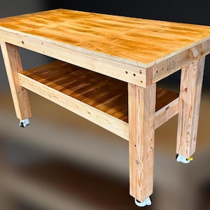 Może przedstawiać: Duży, prostokątny drewniany stół warsztatowy z naturalnym wykończeniem. Stół warsztatowy posiada dolną półkę i jest wsparty na grubych, solidnych nogach. Wyposażony jest w kółka ułatwiające przemieszczanie. Widoczne usłojenie drewna.