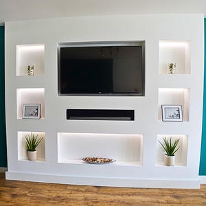 Pode incluir: Um centro de entretenimento branco embutido com uma grande televisão preta, prateleiras embutidas e iluminação integrada. As prateleiras exibem pequenas plantas, fotos emolduradas e itens decorativos. A parede está contra um fundo azul-petróleo.
