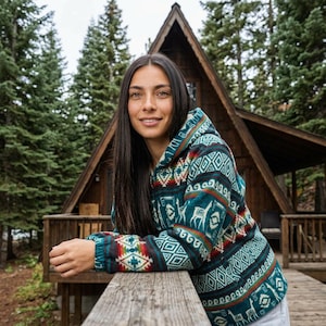 Könnte beinhalten: Eine Kapuzenjacke mit teal-, rot- und weißfarbenem Muster mit geometrischem und Tiermotiv. Die Jacke wird im Freien vor einer Holzhütte und immergrünen Bäumen getragen.