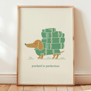 以下が含まれることがあります： 緑色のコートを着て、緑とオレンジ色のキャンプ用ロールを運んでいる漫画のダックスフントが描かれた額入りのプリント。犬の下には「packed to perfection」というテキストがあります。