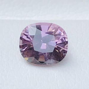 May include: A faceted, oval-shaped gemstone with a light purple hue. The gem is set against a white background, with a dark border at the bottom. The gem's facets reflect light, creating a sparkling effect.