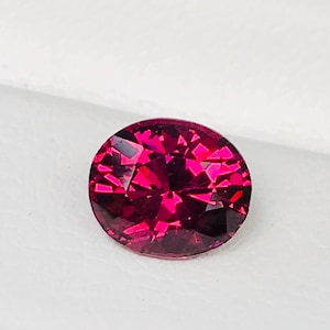 May include: A faceted, oval-cut, deep red gemstone, possibly a ruby or garnet, rests on a white surface within a black jewelry box. The gem's facets catch the light, creating a sparkling effect. The box provides a contrasting backdrop.