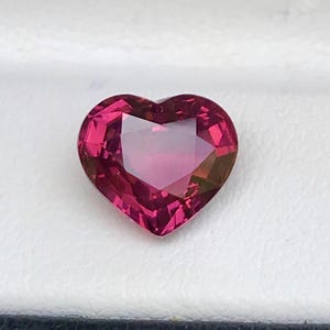 Könnte beinhalten: Ein herzförmiger, facettierter Edelstein in Rosa- und Rottönen. Der Stein ist vor einem weißen Hintergrund in einer schwarz umrandeten Box platziert. Der Schliff des Edelsteins erzeugt mehrere reflektierende Oberflächen.