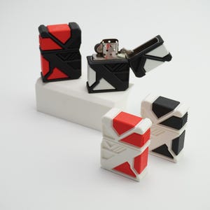 May include: Geometric-patterned lighters in red, black, and white are displayed. One lighter is open, showing the internal mechanism. The lighters are arranged on a white rectangular platform against a white backdrop.