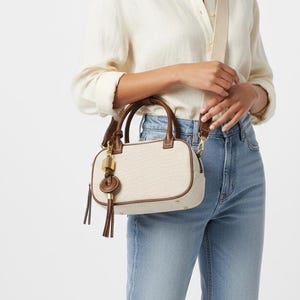 May include: A cream-coloured handbag with brown leather trim and handles. The bag has a detachable shoulder strap and a decorative tassel. The bag is rectangular and appears to be made of canvas material.