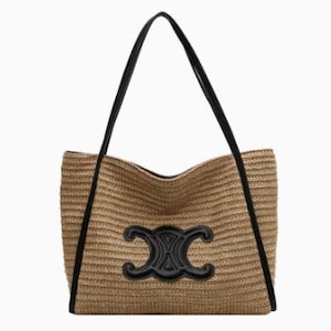 May include: A woven straw tote bag with black leather handles and trim. The bag features a large, black leather emblem on the front. The bag is rectangular and has a spacious interior, perfect for carrying everyday essentials.