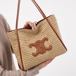 May include: A woven straw tote bag with brown leather trim and handles. The bag features a large brown leather emblem on the front. The bag is held by a person.