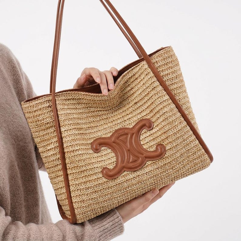 May include: A woven straw tote bag with brown leather trim and handles. The bag features a large brown leather emblem on the front. The bag is held by a person.