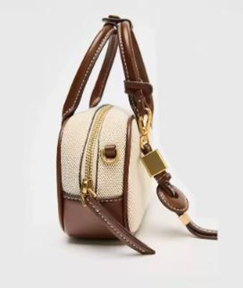 May include: A small, rectangular handbag with a cream-coloured woven body and brown leather trim. It features a gold zip, a short handle, and a detachable shoulder strap. The bag has a structured design and a minimalist aesthetic.