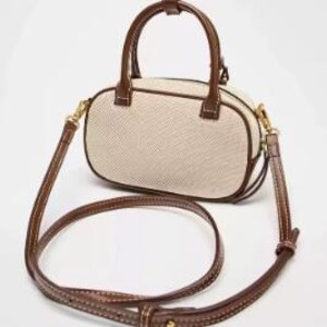 May include: A small, oval handbag with a woven beige body and brown leather trim. It has a top handle and a detachable shoulder strap. The bag features gold-coloured hardware and a stitched design.