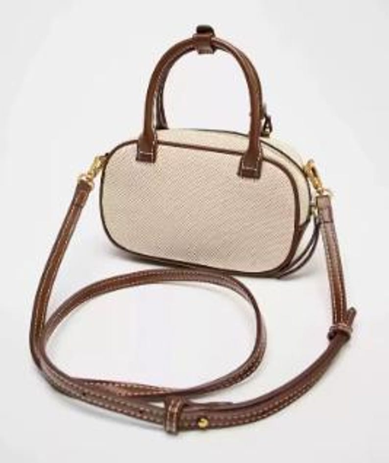 May include: A small, oval handbag with a woven beige body and brown leather trim. It has a top handle and a detachable shoulder strap. The bag features gold-coloured hardware and a stitched design.