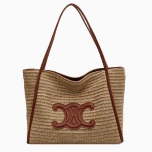 May include: A woven straw tote bag with brown leather handles and trim. The bag features a large, embossed brown leather emblem on the front. The bag is rectangular with a wide opening and long shoulder straps.