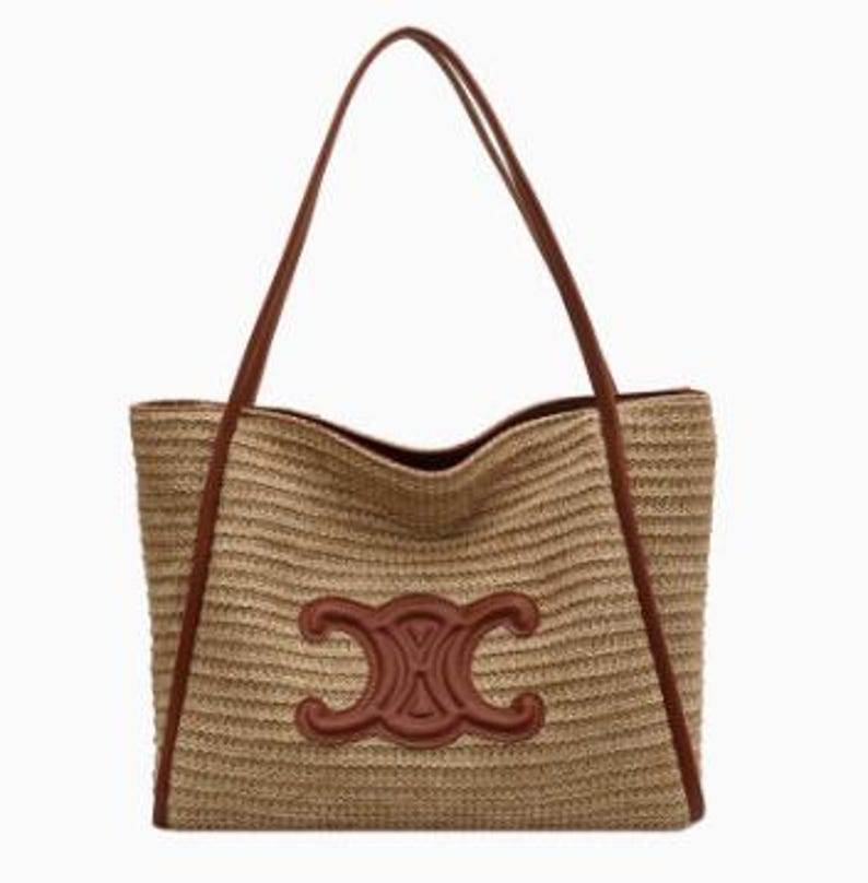 May include: A woven straw tote bag with brown leather handles and trim. The bag features a large, embossed brown leather emblem on the front. The bag is rectangular with a wide opening and long shoulder straps.