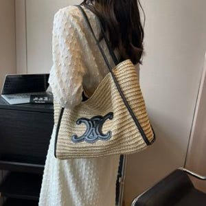 May include: A beige woven tote bag with black leather trim and straps. The bag features a large, dark blue emblem on the front. The bag is carried over the shoulder.