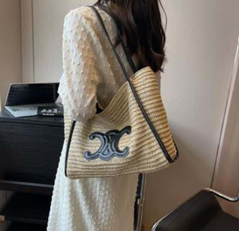 May include: A beige woven tote bag with black leather trim and straps. The bag features a large, dark blue emblem on the front. The bag is carried over the shoulder.