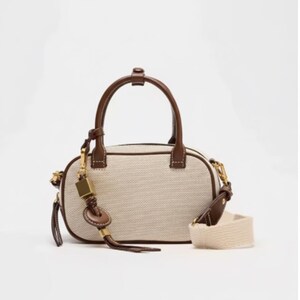 May include: A small, cream-coloured handbag with brown leather trim and handles. The bag features a detachable cream-coloured strap and a gold-toned charm. The bag has a woven texture and a zip closure.